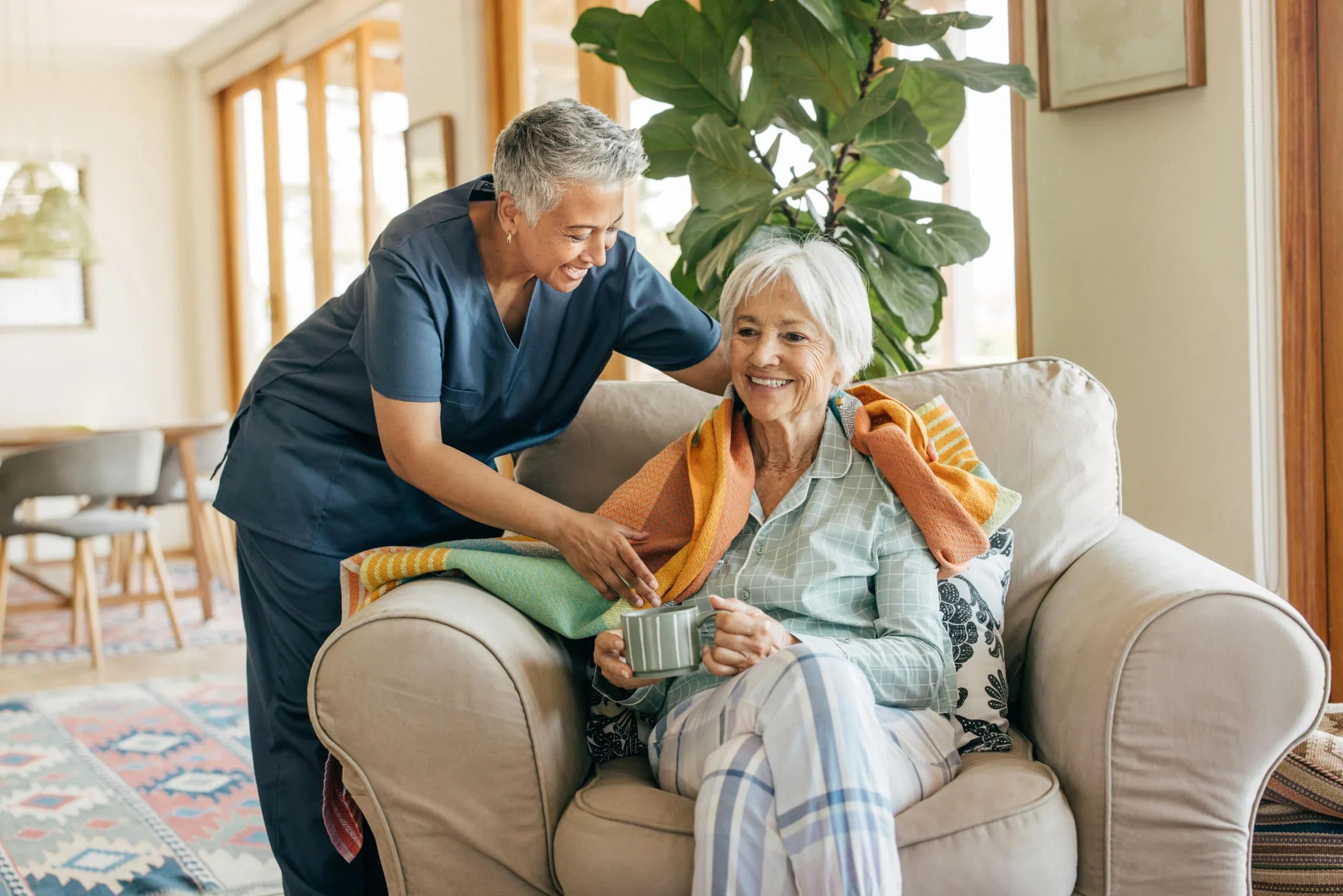 Caregiver placing a colorful blanket on a seated senior woman holding a mug in a cozy living room setting.