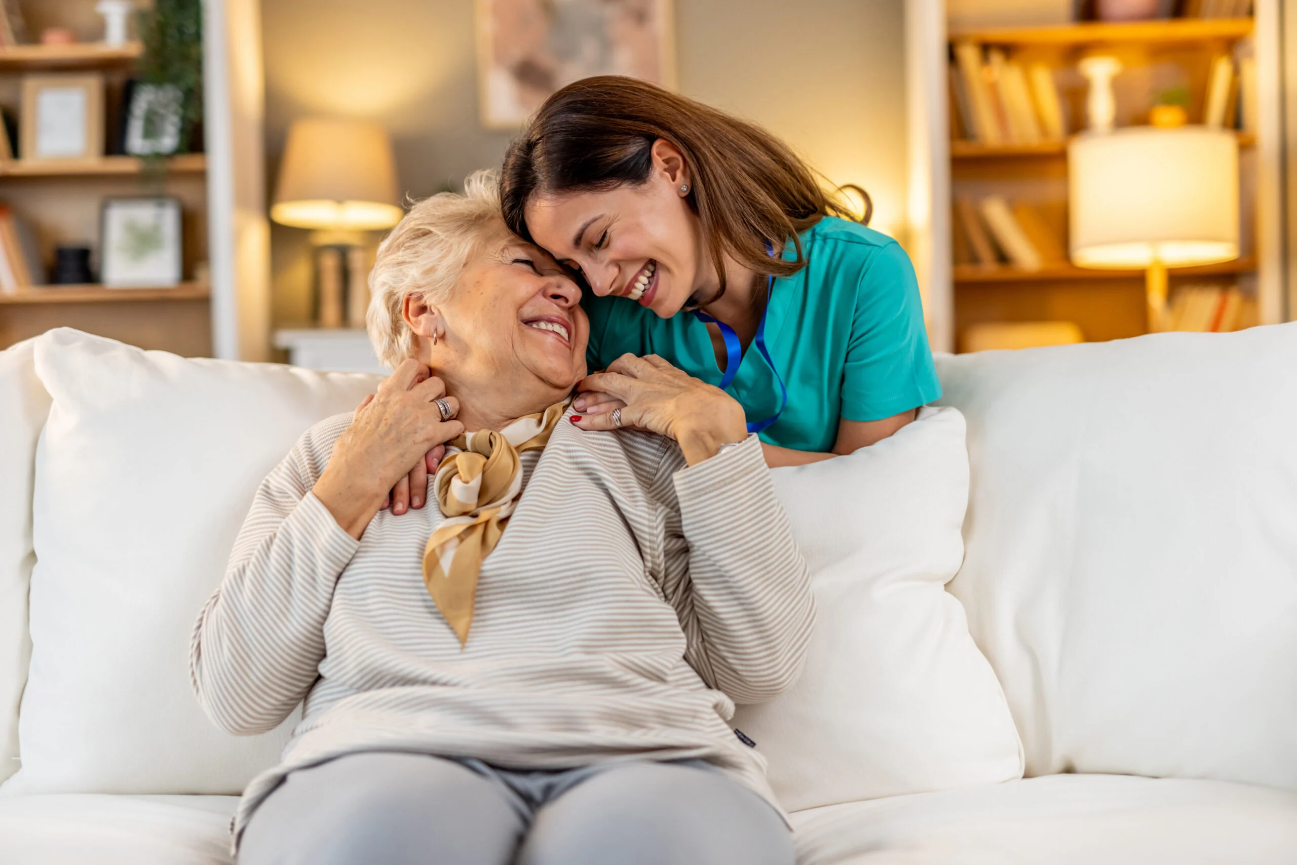Caregiver and senior woman sharing a joyful moment on a living room sofa in a senior living community setting.