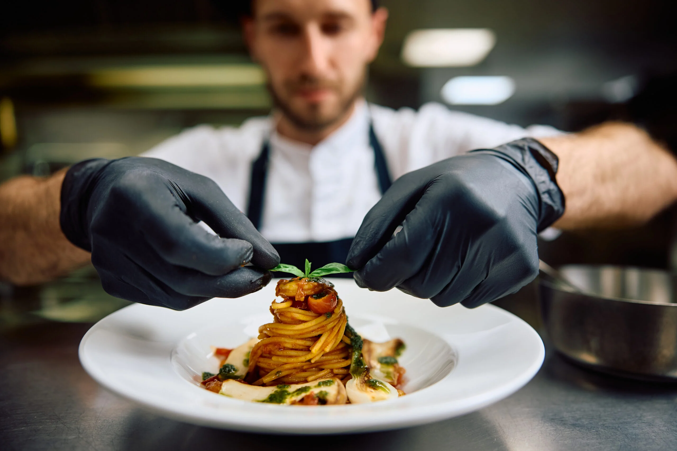 Chef garnishing a plated spaghetti dish with fresh herb in a kitchen.