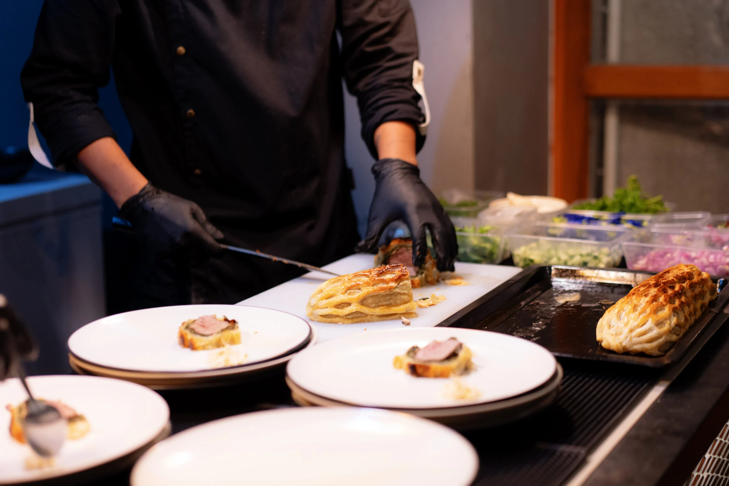 Person wearing black gloves slicing and plating cooked meat dishes with pastry crust on white plates in a kitchen.