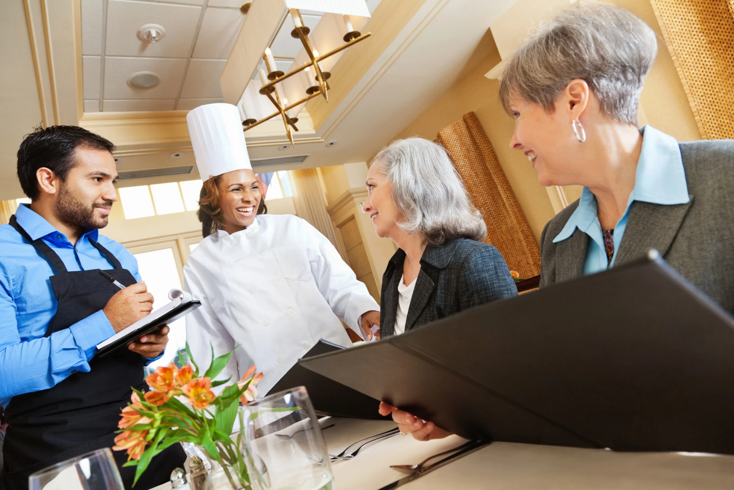 Two older women seated at a dining table interacting with a smiling chef and waiter in a senior living community dining room.