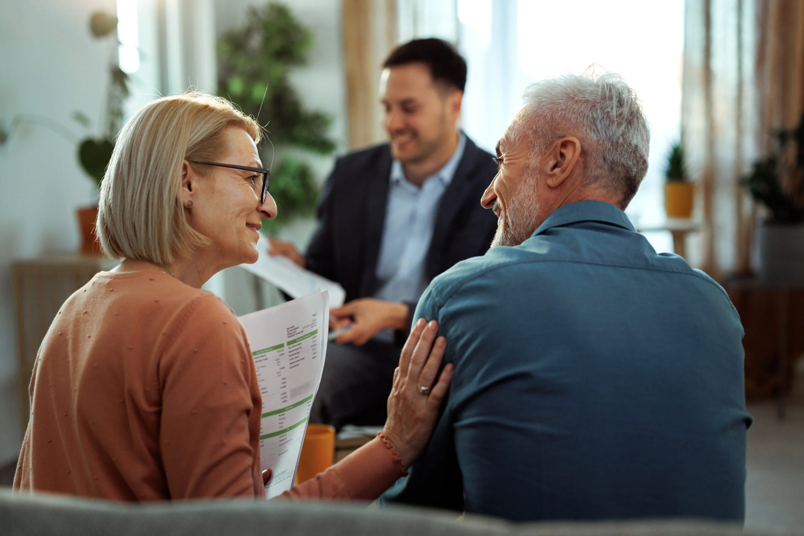 Older couple seated on a couch consulting with a man in a suit holding papers in a bright room with plants.