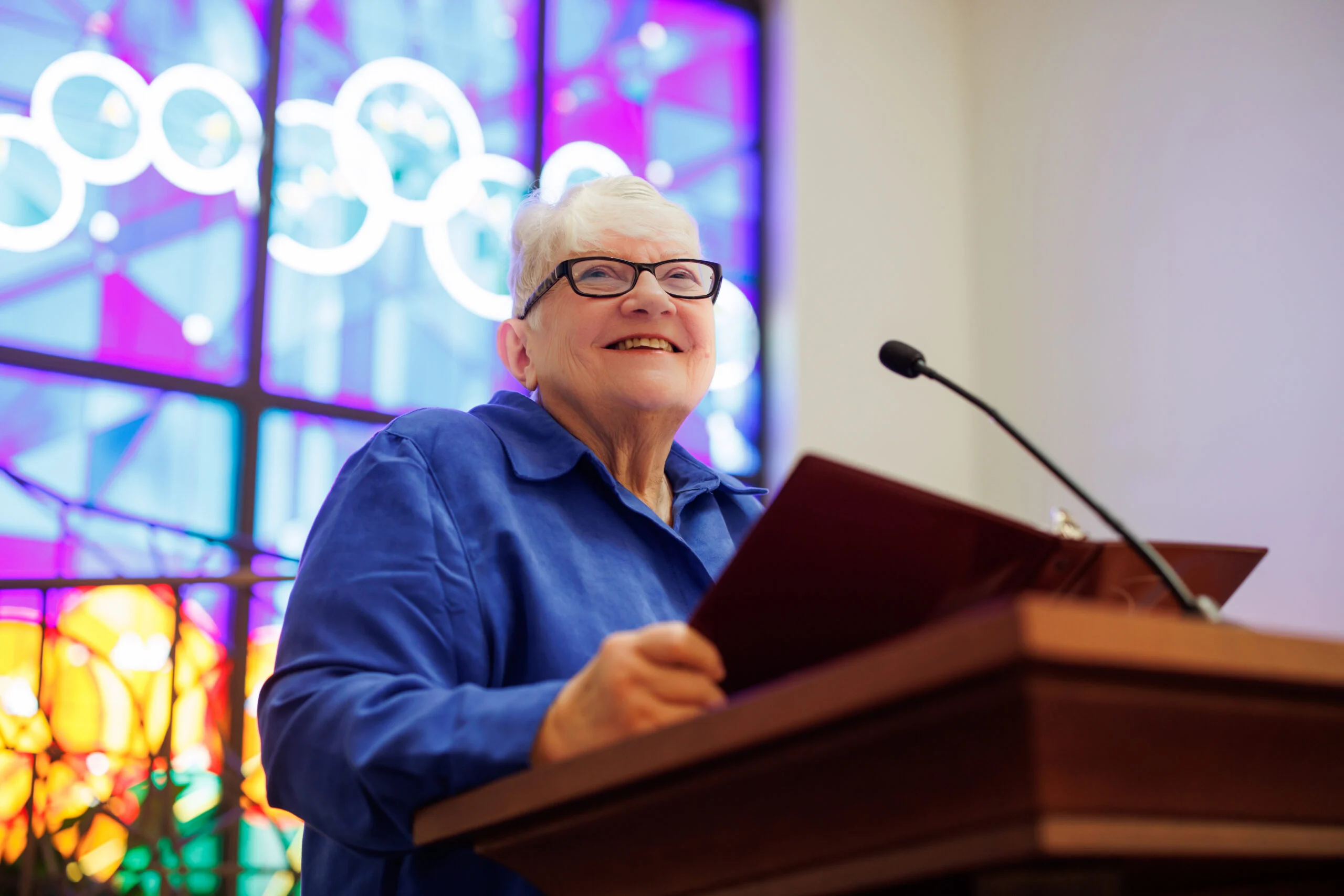 An elderly woman smiling and speaking at a podium with a microphone, holding an open book in front of colorful stained glass windows.
