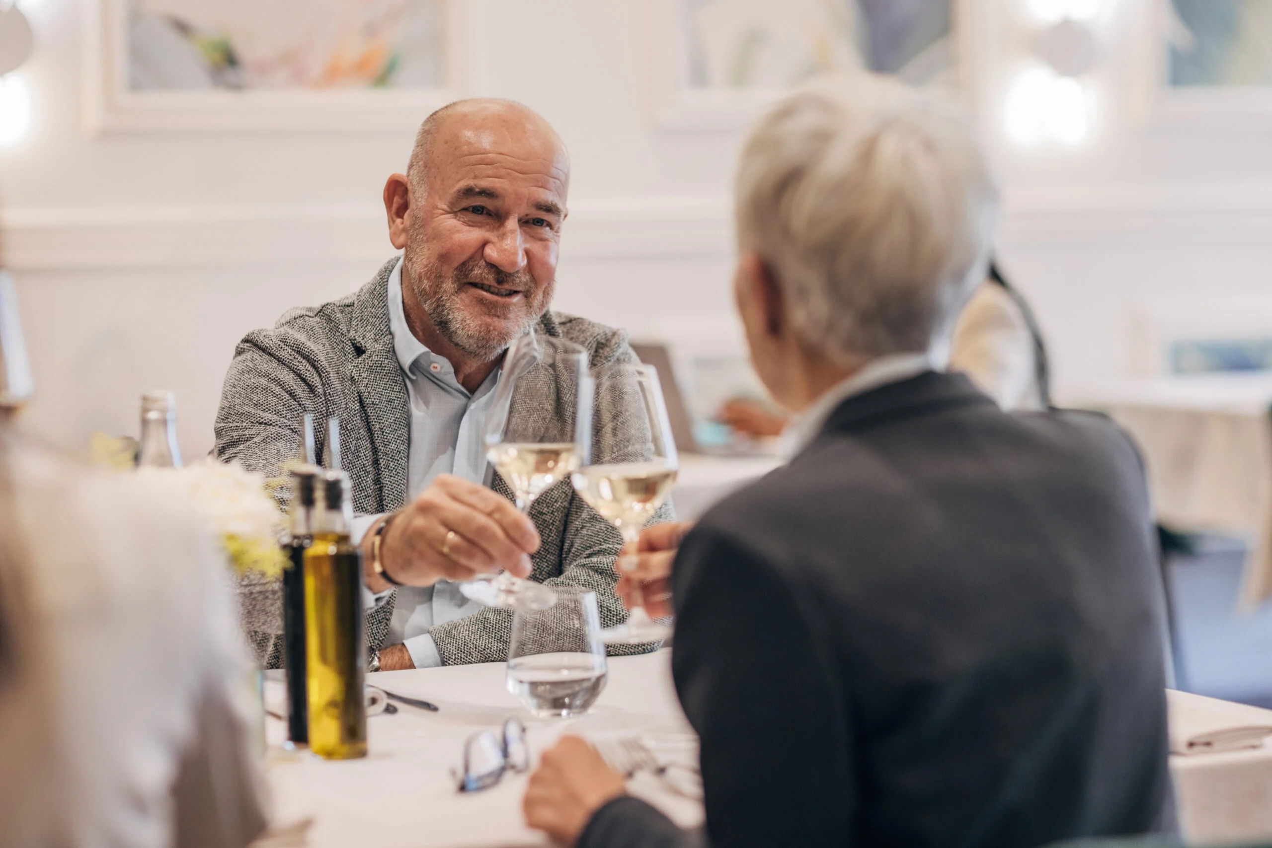 Two seniors at a dining table toasting with wine glasses in a bright elegant dining setting.