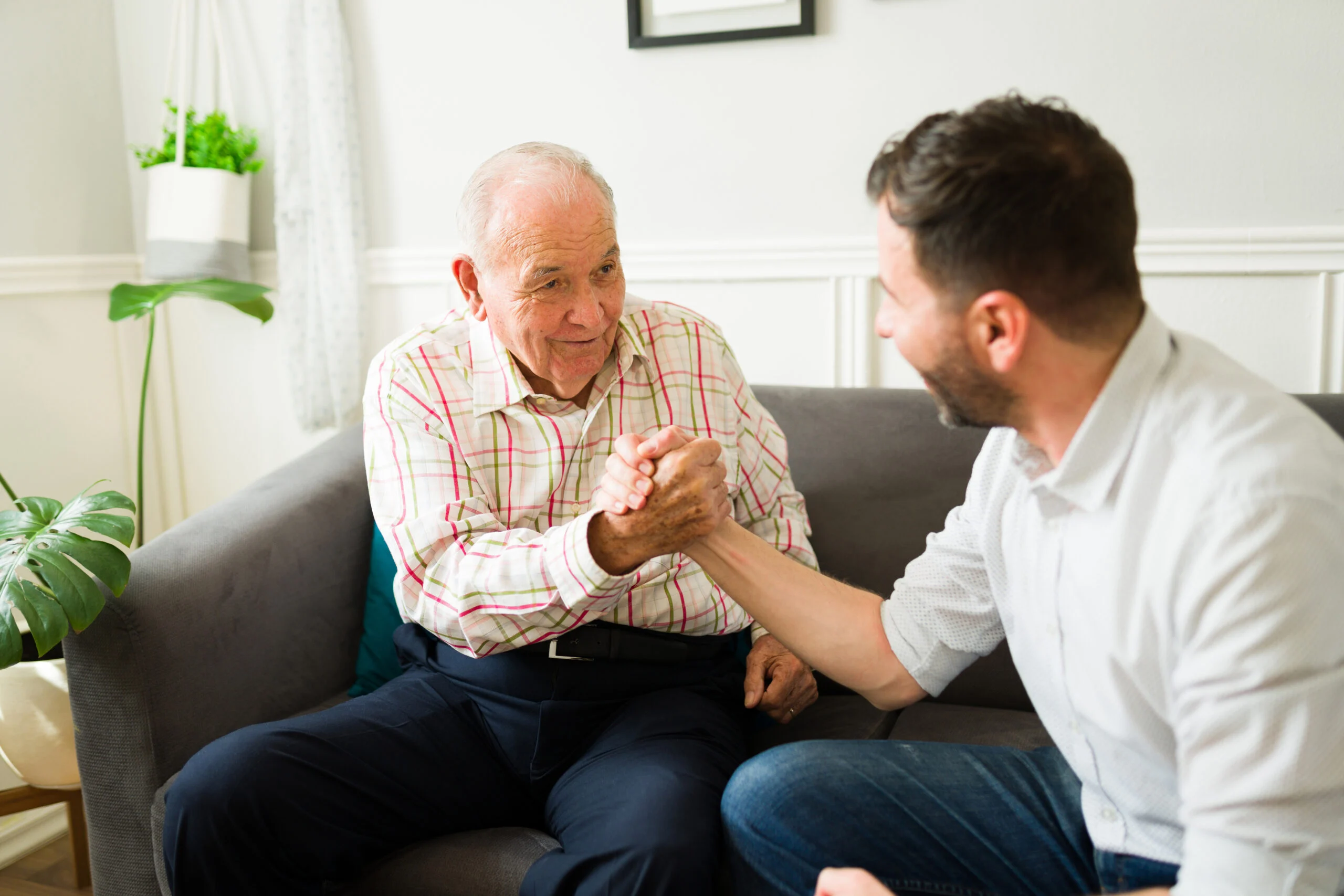 An elderly man and a younger man smiling and shaking hands while seated on a couch in a bright living room.