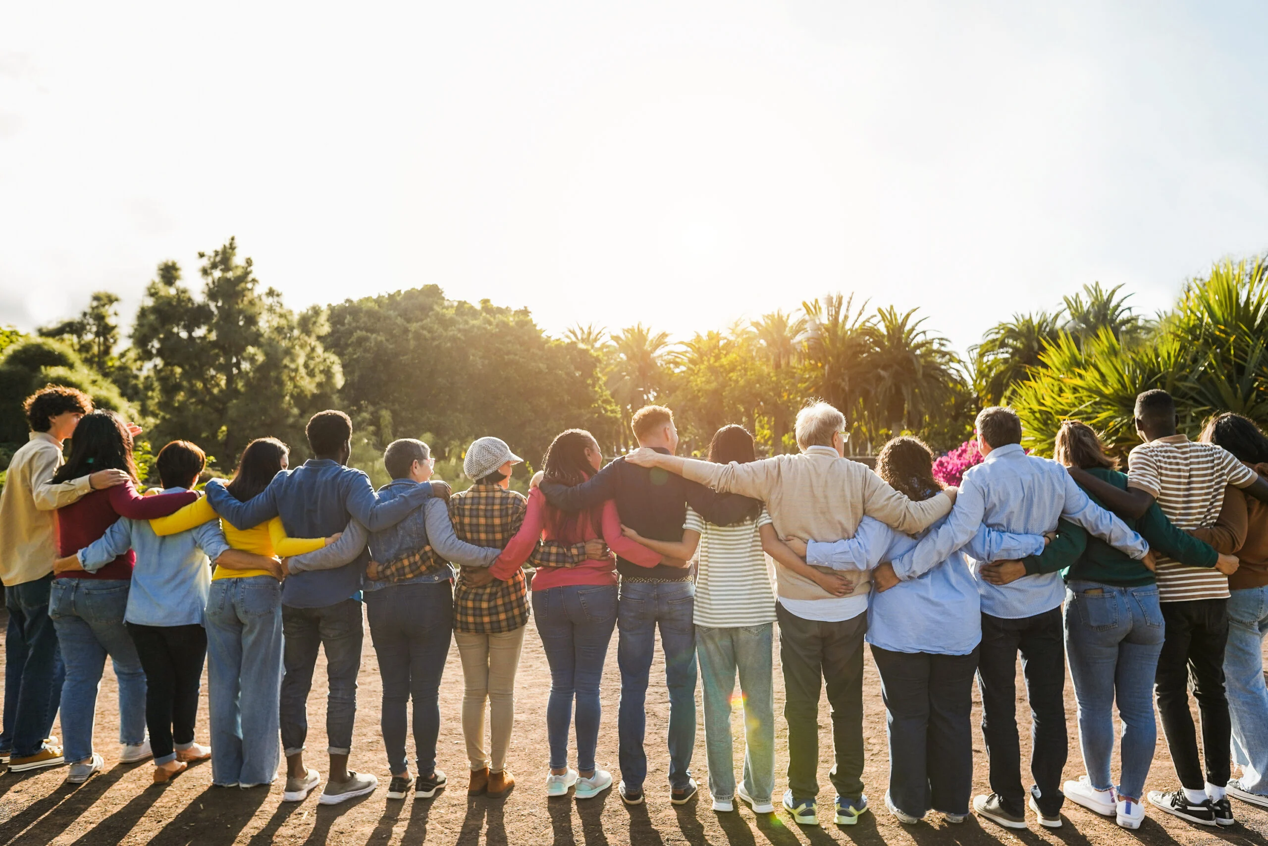 A diverse group standing outdoors with arms around each other’s shoulders in a sunny park setting.