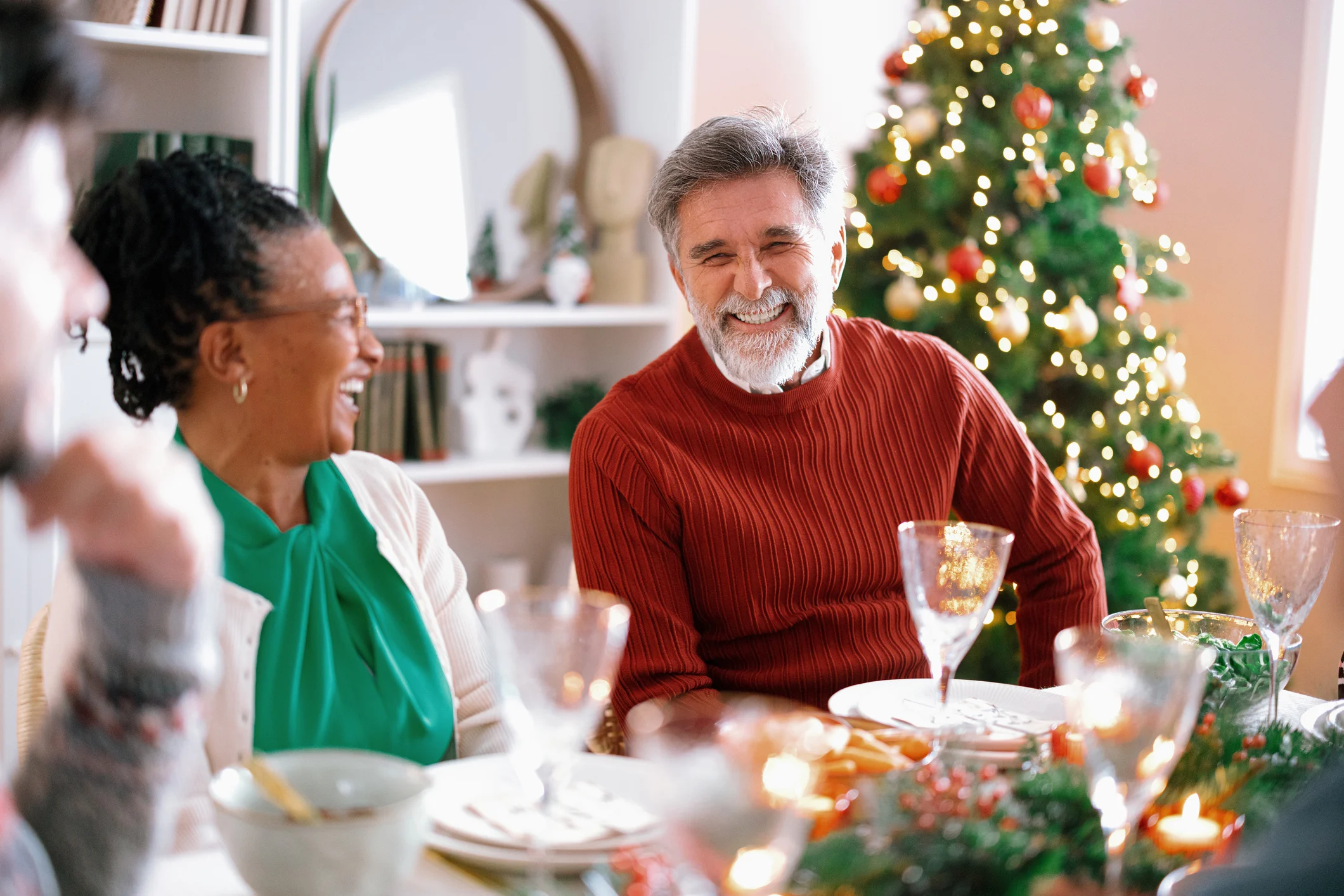 Older adults smiling and socializing around a decorated dining table with a Christmas tree in the background.