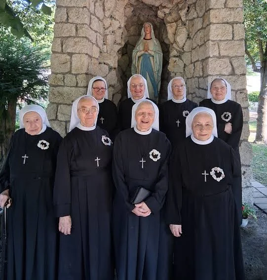 Group of elderly women in nun habits standing outdoors in front of a stone grotto with a Virgin Mary statue.