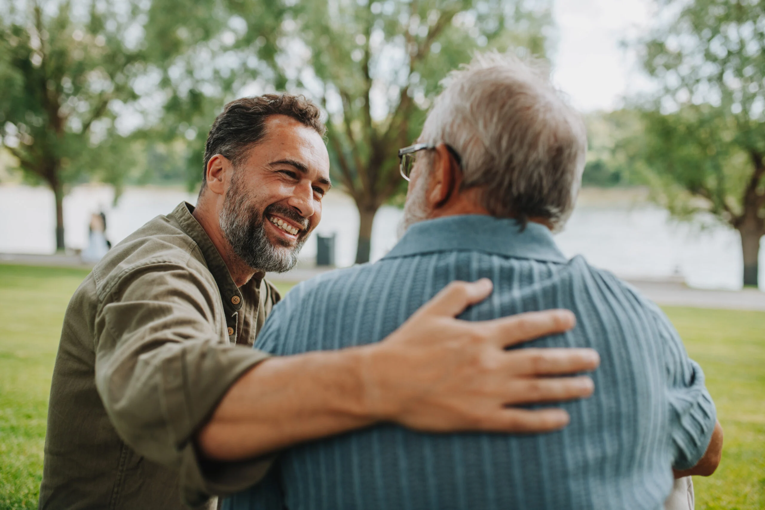 A younger man smiles warmly while embracing an older man outdoors on a grassy area near trees and water.