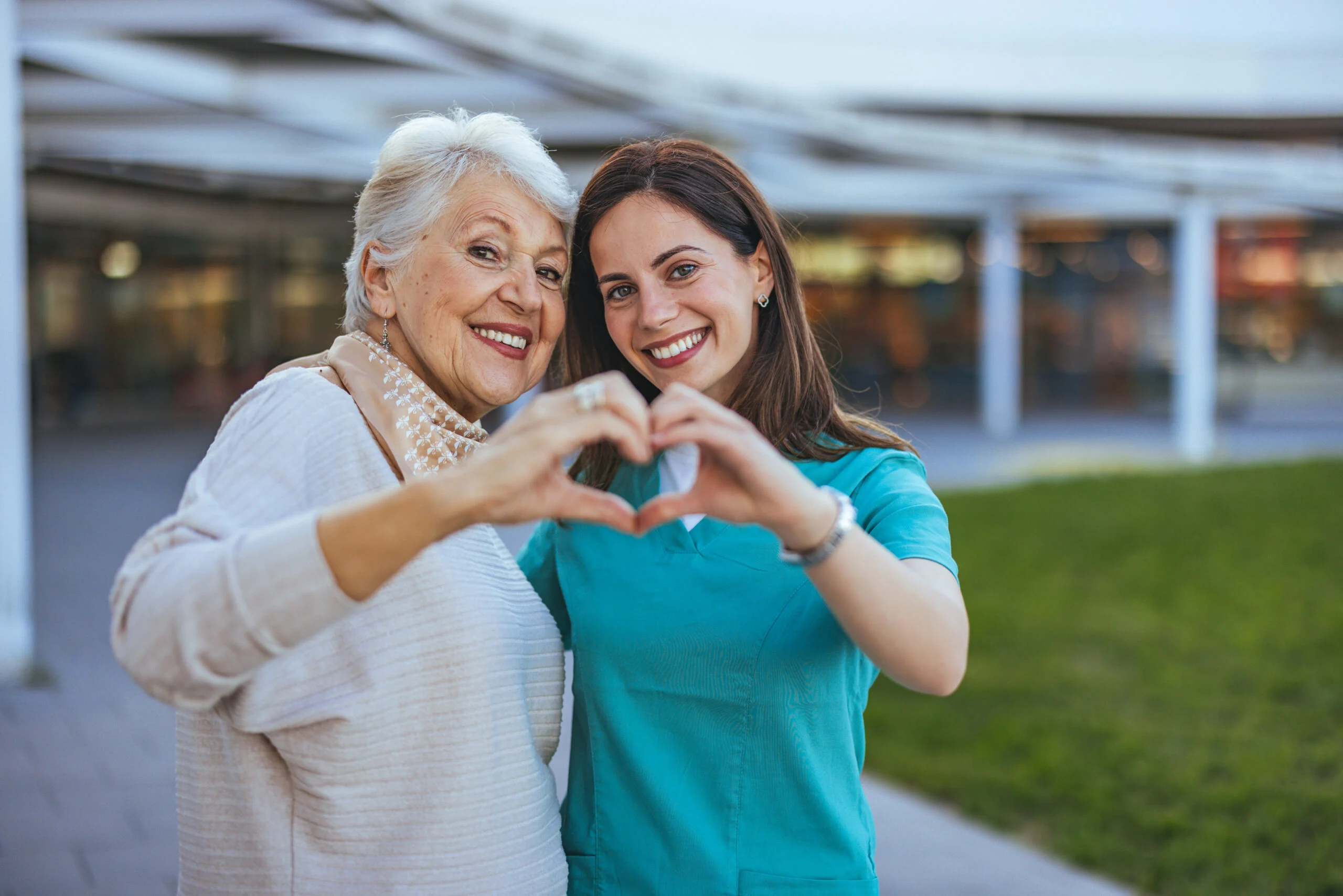 An older woman and a younger caregiver smiling outdoors near a senior living community making a heart shape with their hands.