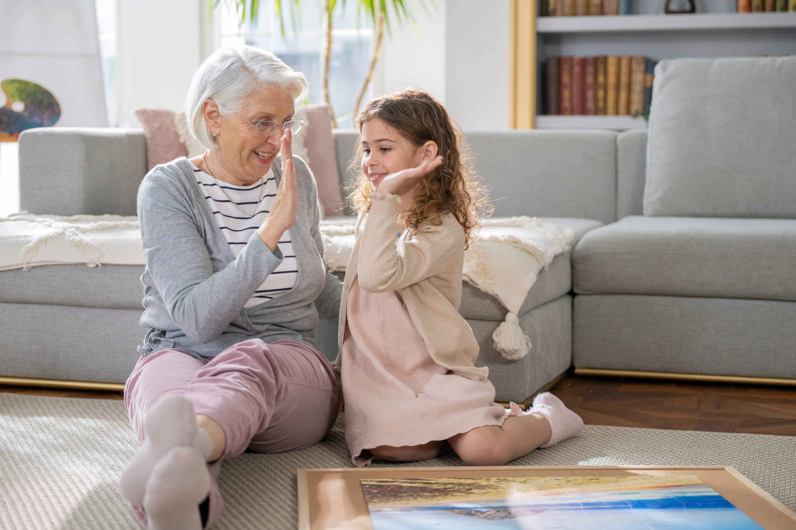 An elderly woman and a young girl sitting on the floor in a living room, giving each other a high-five during an activity.