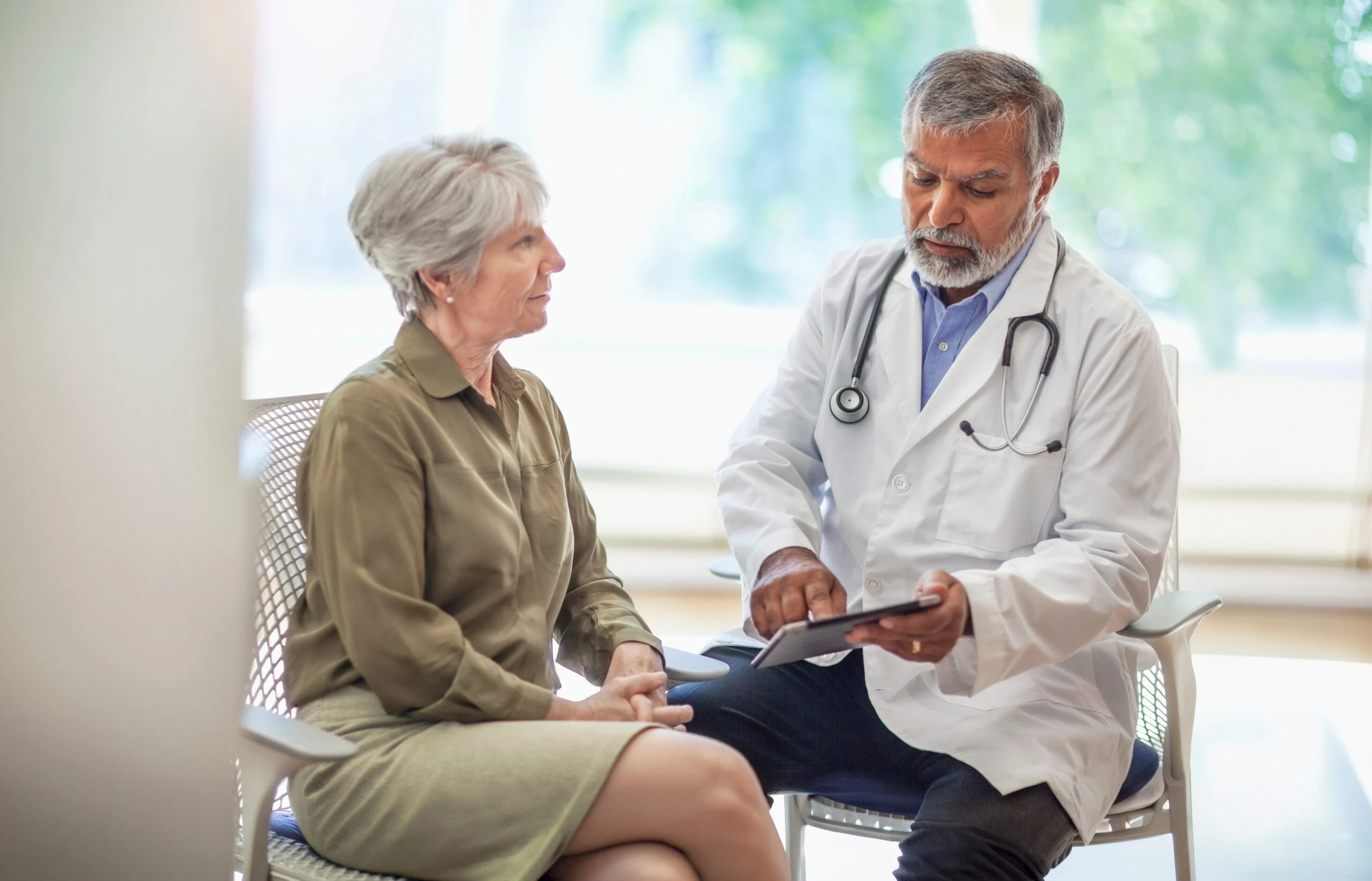 Older adult woman consulting with a healthcare professional who is referencing a digital tablet in a bright room.