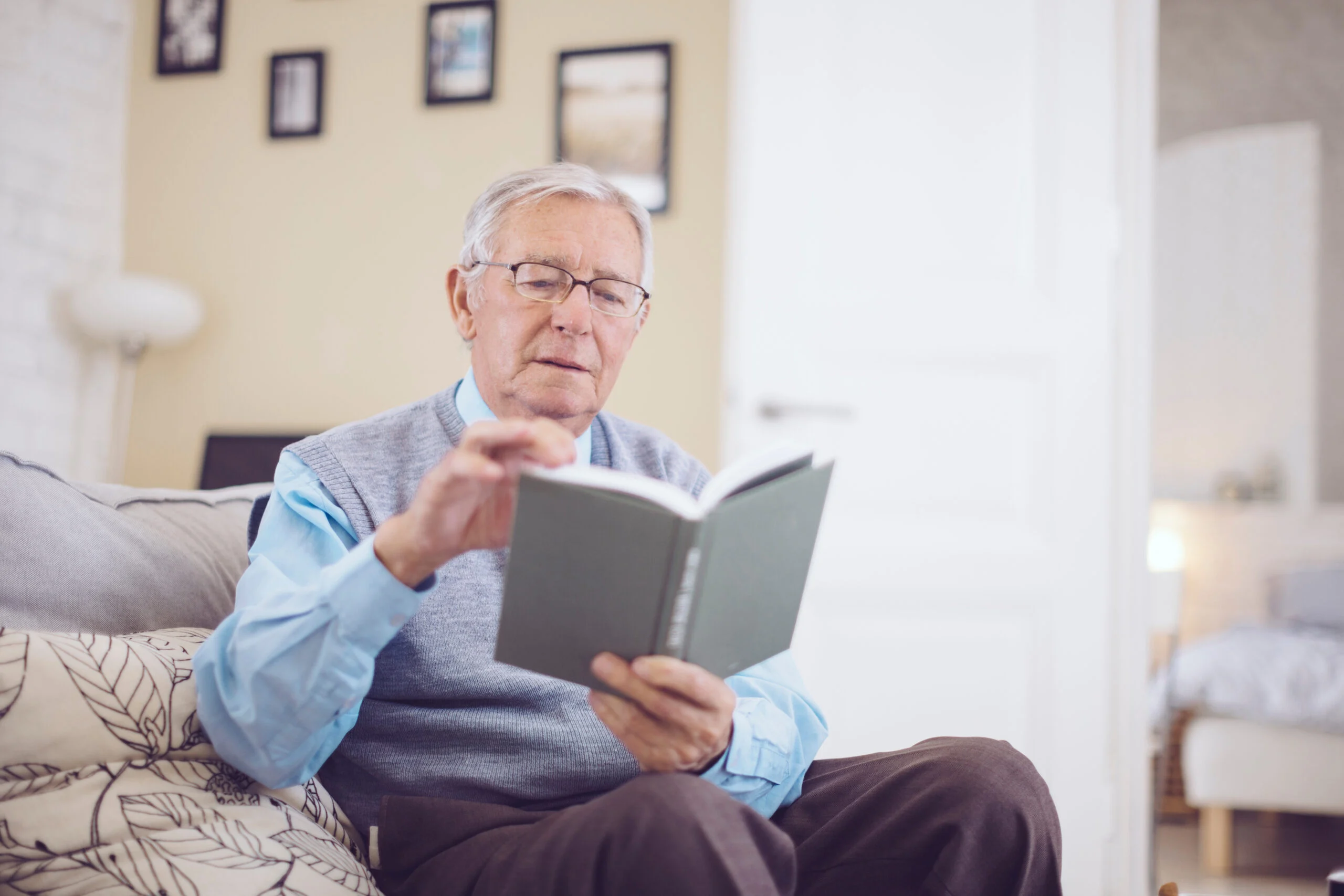 An elderly man reading a book while seated on a couch in a bright living room of a senior living community.