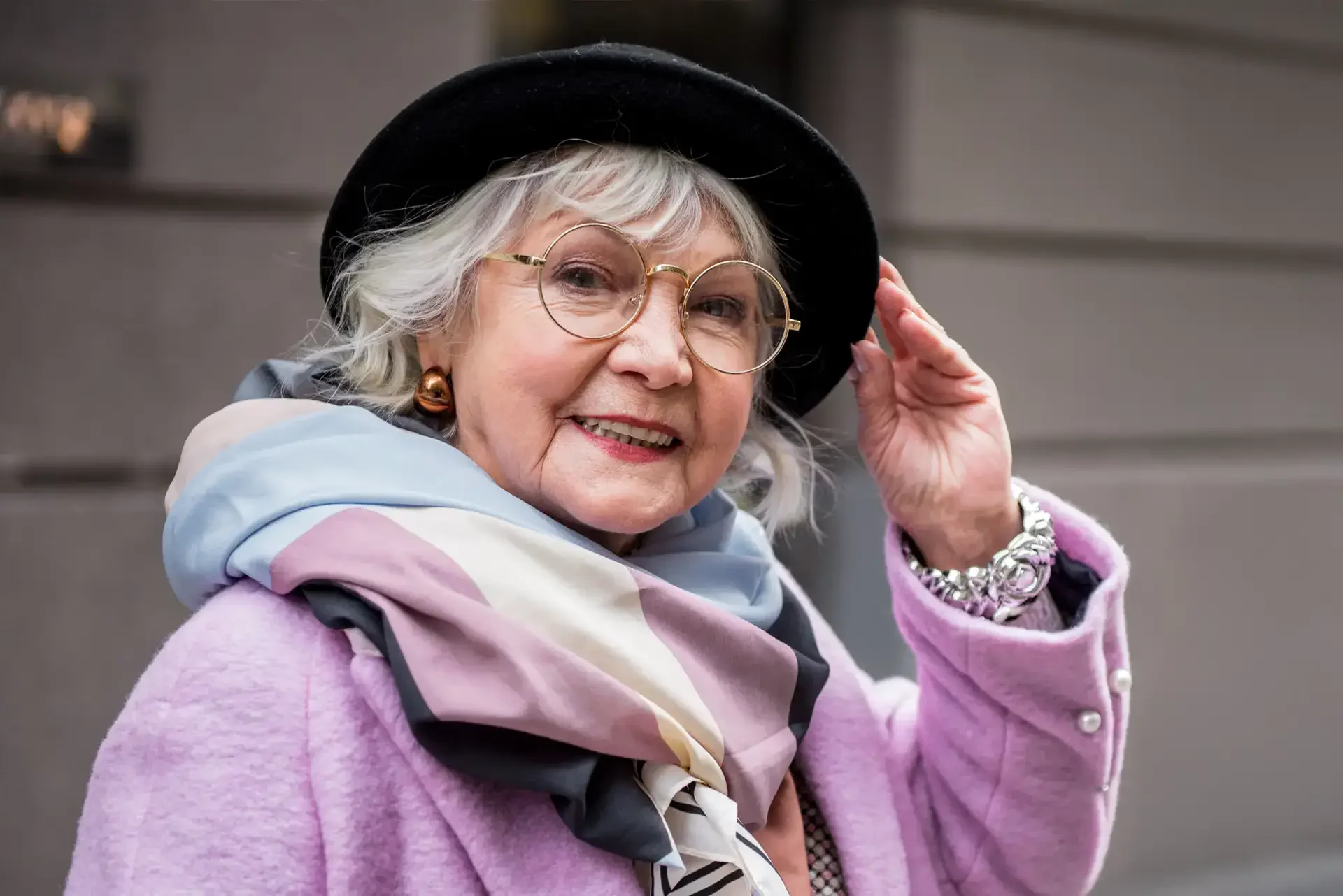 Elderly woman smiling outdoors wearing a pastel purple coat, scarf, glasses, and black hat.