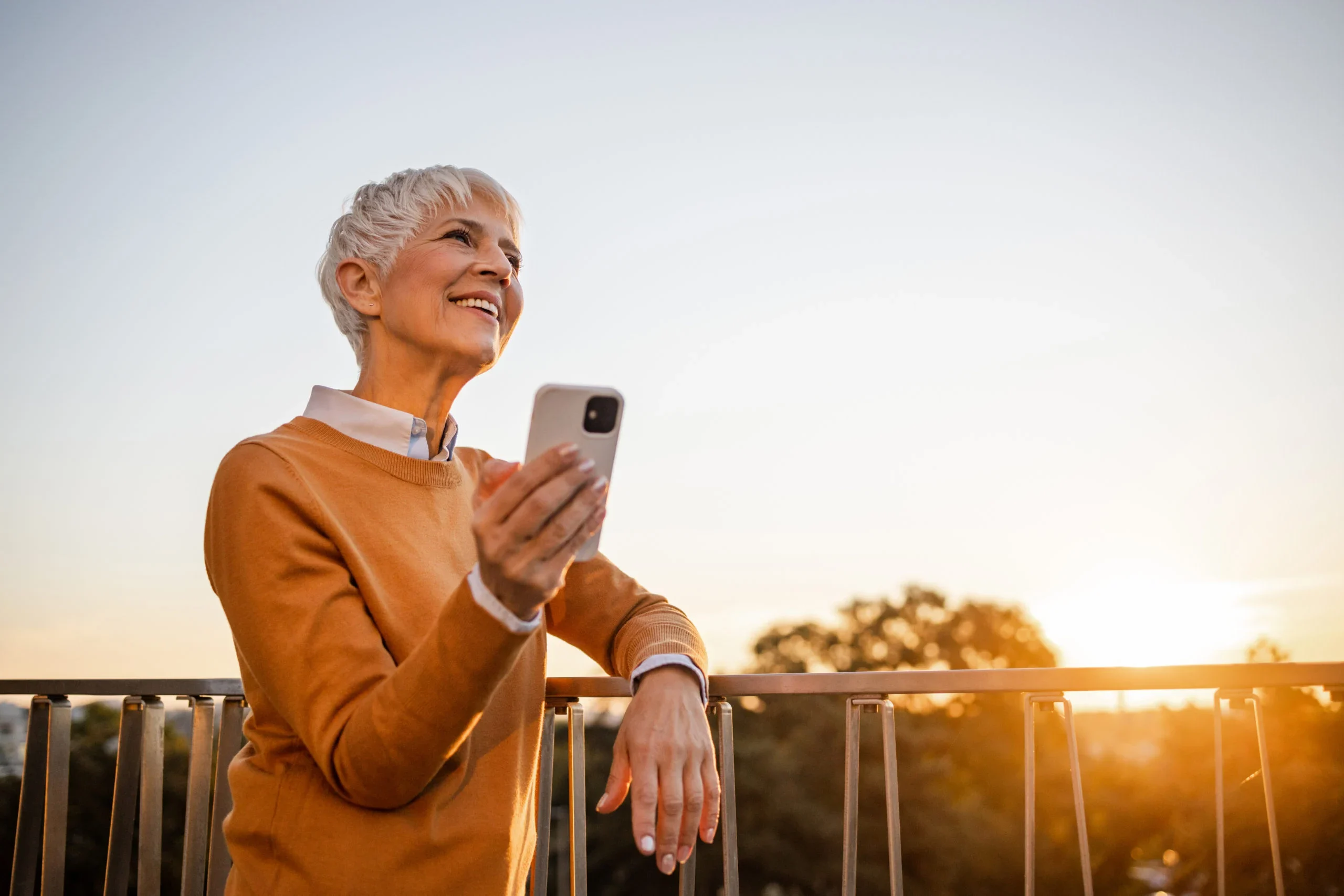 Senior woman standing on a balcony at sunset holding a smartphone and smiling.