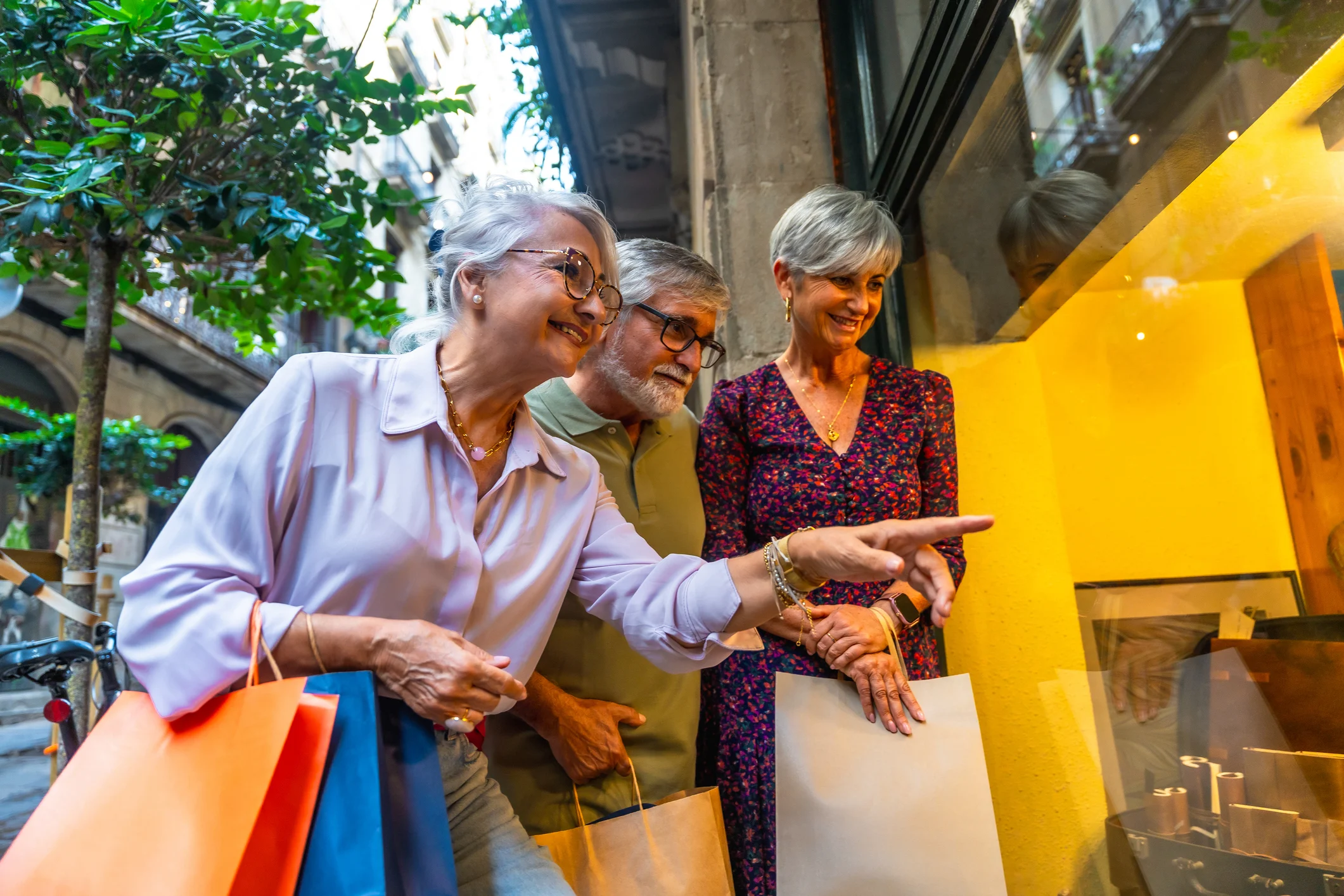 Three senior adults smiling and looking at a store window display while holding shopping bags outdoors in a community setting.