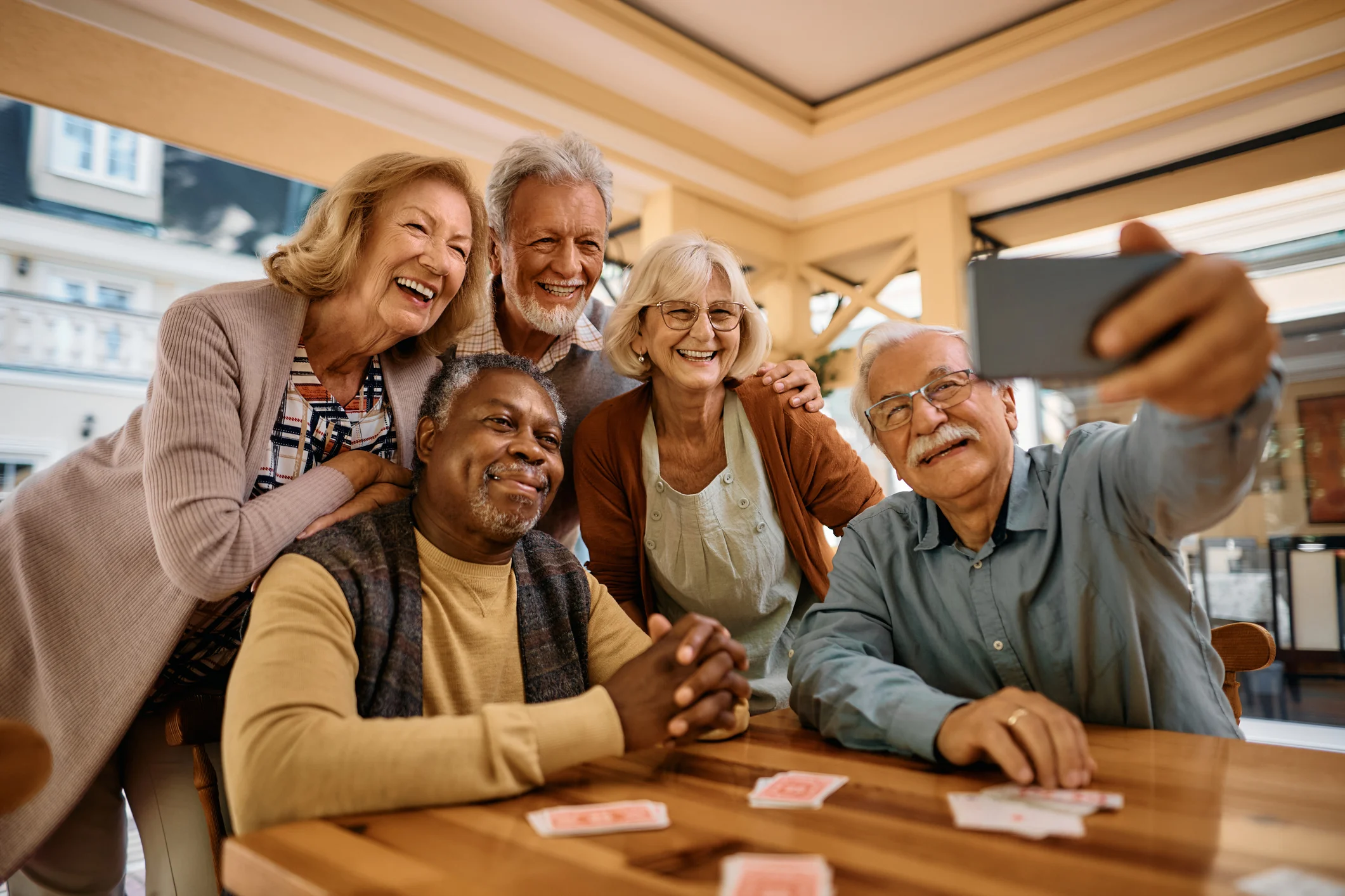 Five seniors gathered around a table with playing cards, smiling and taking a selfie in a warm indoor setting.