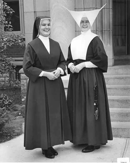 Two persons in traditional religious habits smiling and standing outside near a building entrance.