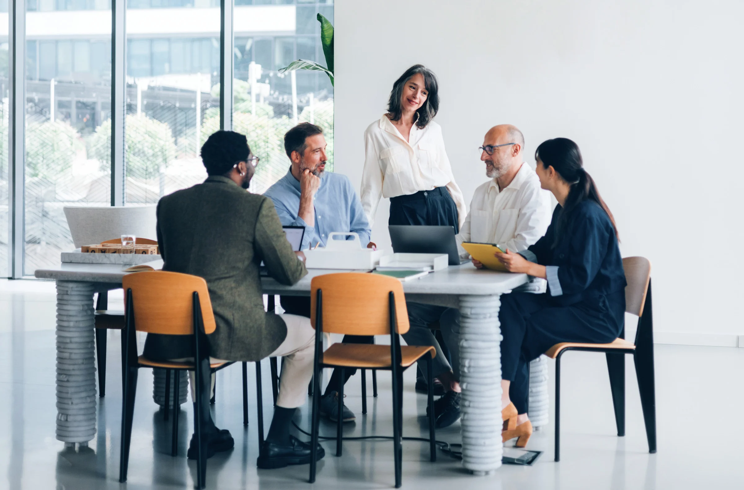 Five professionals gathered around a table in a bright office space engaged in a collaborative meeting with laptops and tablets.