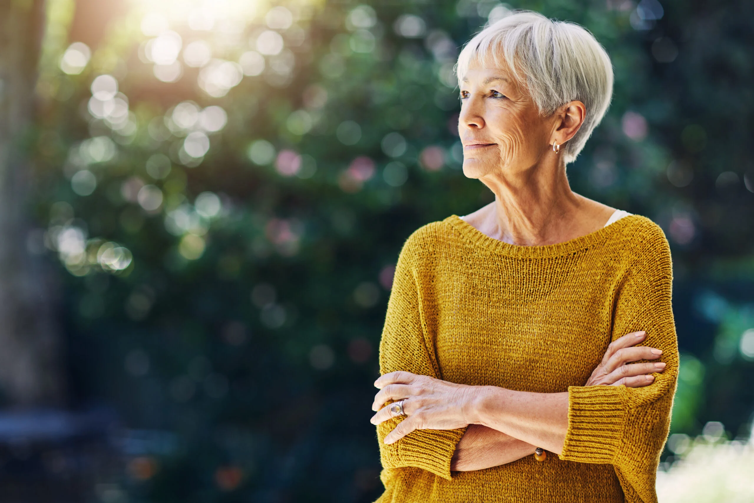 An older woman stands outdoors with arms crossed, looking thoughtfully to the side with a blurred garden background.
